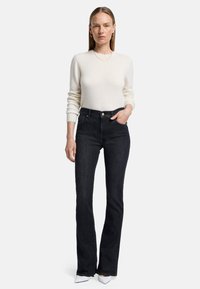 Pull en tricot beige clair à manches longues, associé à un jean flare bleu foncé. Le jean est taille haute et possède des poches avant.