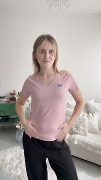 Young woman standing indoors wearing a pink Hugo T-shirt and black pants, with a white couch and green side table in the background.
