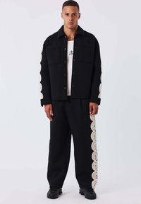 Schwarze Jacke mit zwei Fronttaschen, versehen mit gemustertem Spitzenbesatz an den Ärmeln; kombiniert mit schwarzen Hosen mit passenden Spitzenakzenten. Schwarze Sneakers.