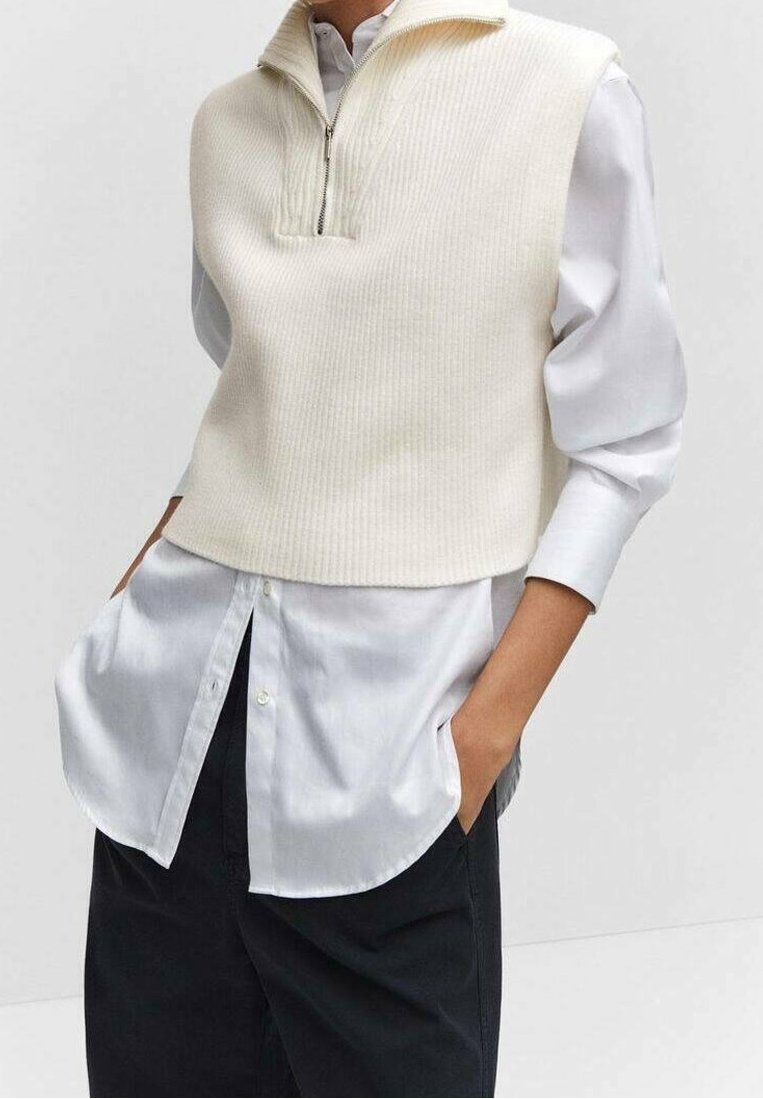 Crème kleurige geribbelde mouwloze trui met een hoge hals en ritssluiting, gedragen over een witte blouse met knopen en donkere broek.