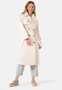 Manteau trench beige en tissu léger, présentant un design croisé, de grands boutons, une taille ceinturée et des poches latérales.