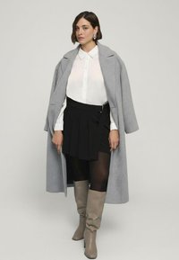 Grau wolliger Mantel über einem weißen Button-up-Hemd und einem schwarzen, gefältelten Minirock. Beigefarbene kniehohe Stiefel runden das Outfit ab und setzen einen strukturierten Akzent.