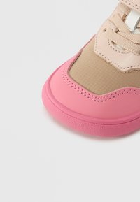 Αθλητικό παπούτσι με ροζ ελαστική σόλα, μπεζ επάνω μέρος από δίχτυ και δερμάτινες λεπτομέρειες. Στρογγυλεμένο μπροστινό μέρος με αντίθετες υφές και ραμμένα διακοσμητικά στοιχεία.