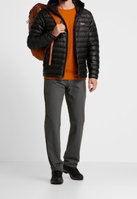 Mężczyzna w czarnej kurtce puchowej, pomarańczowej koszulce, szarych spodniach i czarnych butach, noszący brązowy plecak na jednym ramieniu na białym tle.