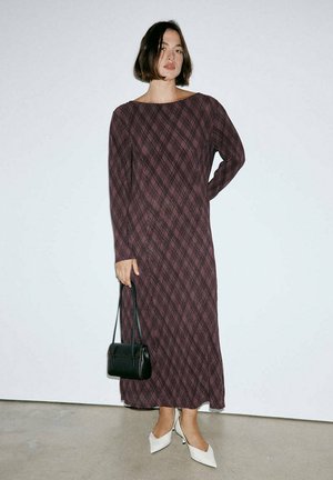 Femme portant une robe à carreaux violette, manches longues, longueur aux chevilles, tenant un petit sac noir, chaussée de talons blancs à bout pointu, debout contre un mur uni.