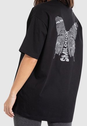 Osoba ubrana w czarny luźny t-shirt z białym wzorem linii papilarnych i napisem "SMILE000X" na plecach, na jednolitym jasnym tle.