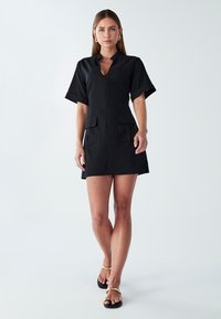 Črna obleka s kratkimi rokavi in stoječim ovratnikom, ki ima ključavnico na dekolteju in dve sprednji žepek. Gladek material, minimalističen dizajn.