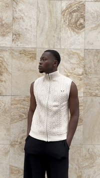 Cream-colored, sleeveless cable knit sweater with a high collar and front zipper. Textured pattern and ribbed hem, worn over black pants.