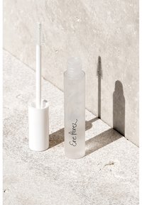 Klarer Behälter mit weißem Wimpernserum-Applikator, der auf einer hellen, strukturierten Oberfläche liegt und Schatten an eine Steinmauer wirft.