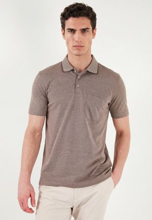 Mand med kort mørkt hår iført en taupe kortærmet poloshirt og lysebeige bukser, stående mod en ensfarvet lys baggrund.