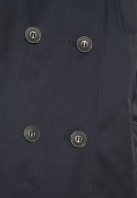 Tejido oscuro con cuatro grandes botones oscuros dispuestos en dos pares verticales paralelos y ojetes de botones cosidos visibles.