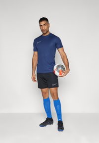 Mørkeblå sportsbluse med korte ærmer, sorte shorts, blå sokker og sorte fodboldstøvler. Holder en mønstret fodbold.