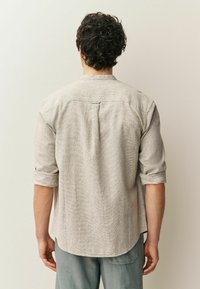 Camicia grigio chiaro con trama, collo con bottoni, maniche arrotolate e orlo posteriore allungato. Design semplice con motivi sottili.