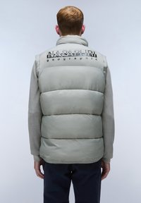 Ljust grå dunstväst med hög krage, logotryck på baksidan, slät yta och elastisk nederkant. Buren över en beige långärmad tröja.
