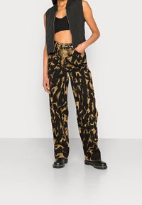 Schwarze gesteppte Weste über einem schwarzen Croptop, kombiniert mit hochgeschnittenen schwarzen Hosen mit einem goldenen abstrakten Muster und schwarzen Stiefeln.