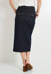 Falda de mezclilla en azul oscuro, hasta la rodilla y con corte recto. Presenta bolsillos traseros bordados y costuras de contraste. Combinada con zapatos de plataforma en color marrón claro.