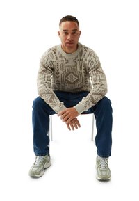 Mann sitzt auf einem Stuhl, trägt einen beigen, gemusterten Pullover, blaue Jeans und hellgrüne Sneaker, hat die Hände gefaltet und schaut nach vorne.