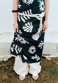 Schwarzes Kleid aus Stoff mit weißen Blumenmustern, mit einem weißen Gürtel gebunden. Kombiniert mit weiten beigen Hosen und flachen Sandalen. Glatte Textur, fließendes Design.