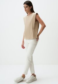 Beżowy bezrękawnik z boczną kieszenią, zestawiony z białymi zwężającymi się dżinsami i białymi trampkami z złotym akcentem. Gładka struktura materiału.