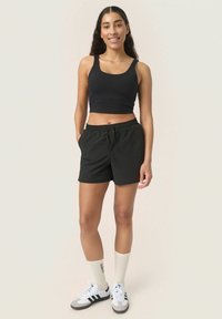 Melnā croptop krekls ar platām siksnām, apvienots ar melniem īsiem šortiem, kuros ir elastīga josta. Baltas zeķes un pelēki sporta apavi noslēdz apģērbu.