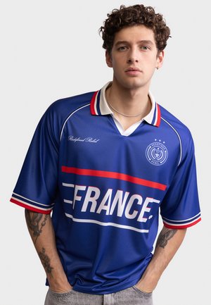 Ung mand med krøllet hår iført en blå sportsjakke med teksten "FRANCE", røde og hvide striber samt tatoveringer på begge underarme.
