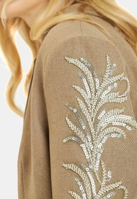 Tejido marrón texturizado con intrincados adornos de cuentas y lentejuelas plateadas y blancas en un patrón de hojas a lo largo de la manga.