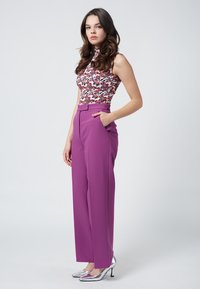 Lila vida byxor med sidofickor som matchas med en blommig ärmlös topp. Modellen står med en hand i fickan och bär silverfärgade klackar.