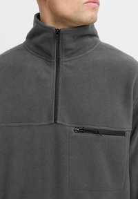 Hallhalli fleece-pullover, millel on kõrge krae, pooltõmblukk ja lukuga rindade tasku. Pehme tekstuur, mugav lõige ja minimaalne disain.