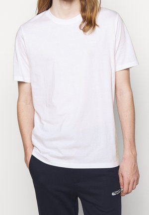 Personne portant un t-shirt blanc uni à manches courtes et un pantalon sombre, avec une main dans la poche, sur un fond clair uni.