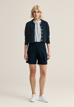 Lyshåret kvinde iført en navyblå cardigan med knapper, hvid mønstret bluse, navy plisserede shorts og hvide sneakers, stående mod en ensfarvet baggrund.