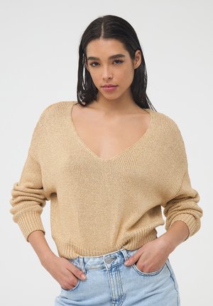 Maglione beige lavorato a maglia con scollo a V profondo, vestibilità ampia e polsini a coste. Abbinato a pantaloncini di jeans azzurri, mani sui fianchi.