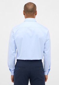 Camicetta azzurra chiara con maniche lunghe e polsini con bottone, abbinata a pantaloni scuri color navy con due tasche posteriori. Tessuto dalla texture liscia.