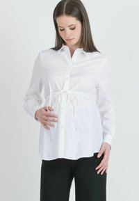 Camicia bianca con bottoni e cintura in vita, dotata di colletto classico e maniche lunghe. Abbinata a pantaloni verde scuro.
