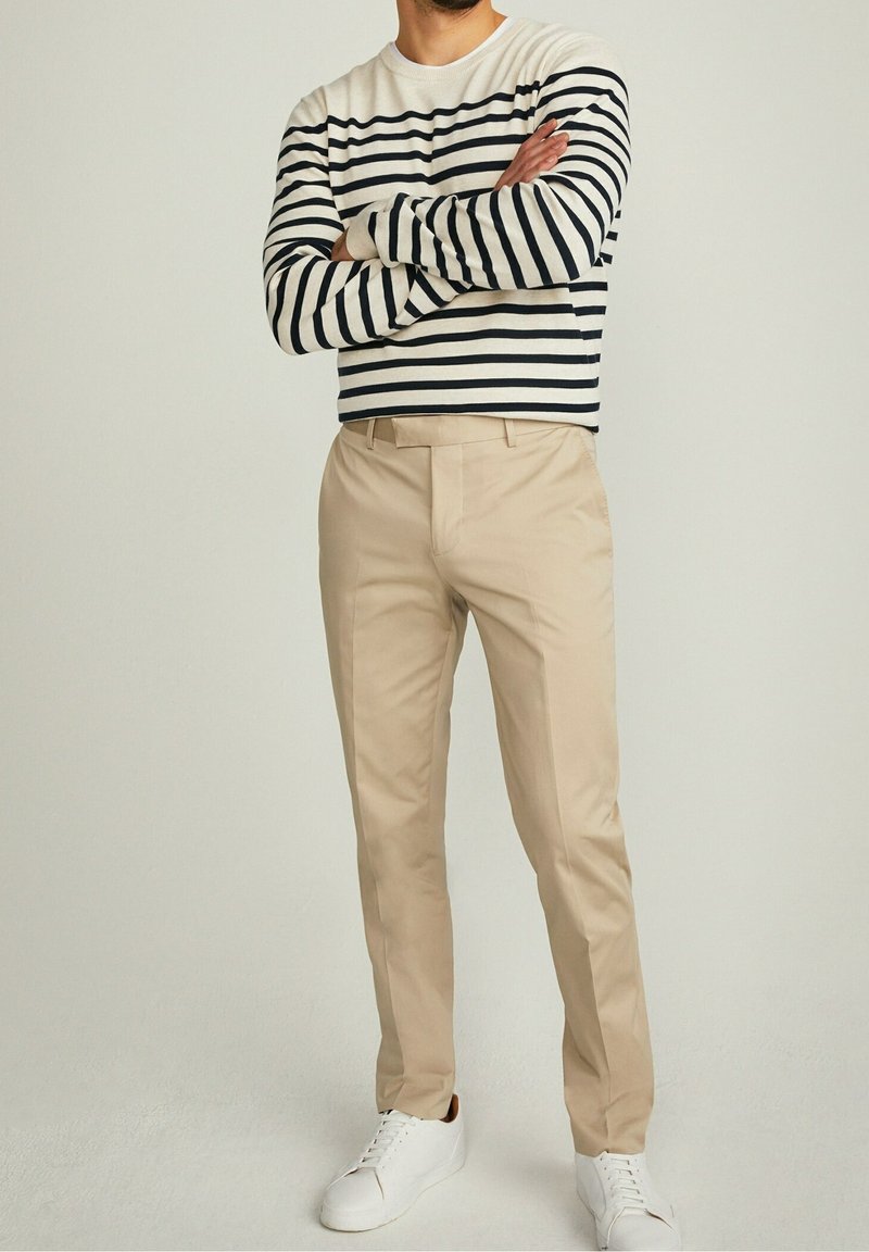 Maglione di cotone a righe in bianco e blu navy, abbinato a pantaloni beige e sneakers bianche. I pantaloni hanno una vestibilità classica con tasche.