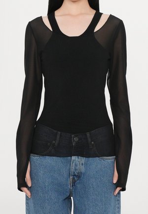 Femme portant un top noir transparent à manches longues superposé sous un top noir sans manches, associé à un jean bleu taille haute.