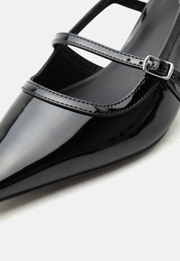 Chaussure plate en cuir verni noir à bout pointu avec deux fines brides réglables et boucles argentées.