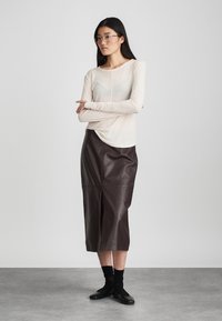 Beżowa bluzka z długim rękawem o miękkiej fakturze, w połączeniu z brązową skórzaną spódnicą midi z detalami szwów i rozcięciem z przodu. Czarne obuwie.