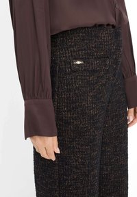 Sorte, teksturerede bukser med guldprikker, der har en dekorativ guld hardwareaccent. Sammen med en mørkebrun silkebluse med puffede ærmer.