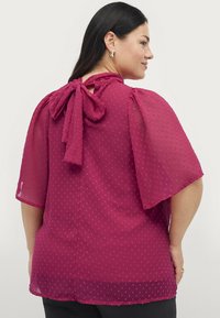 Bordeaux bluse med gennemsigtig, prikket stof; korte puffærmer; knyttet nøglehul detalje bagpå; glat tekstur med let drapering.