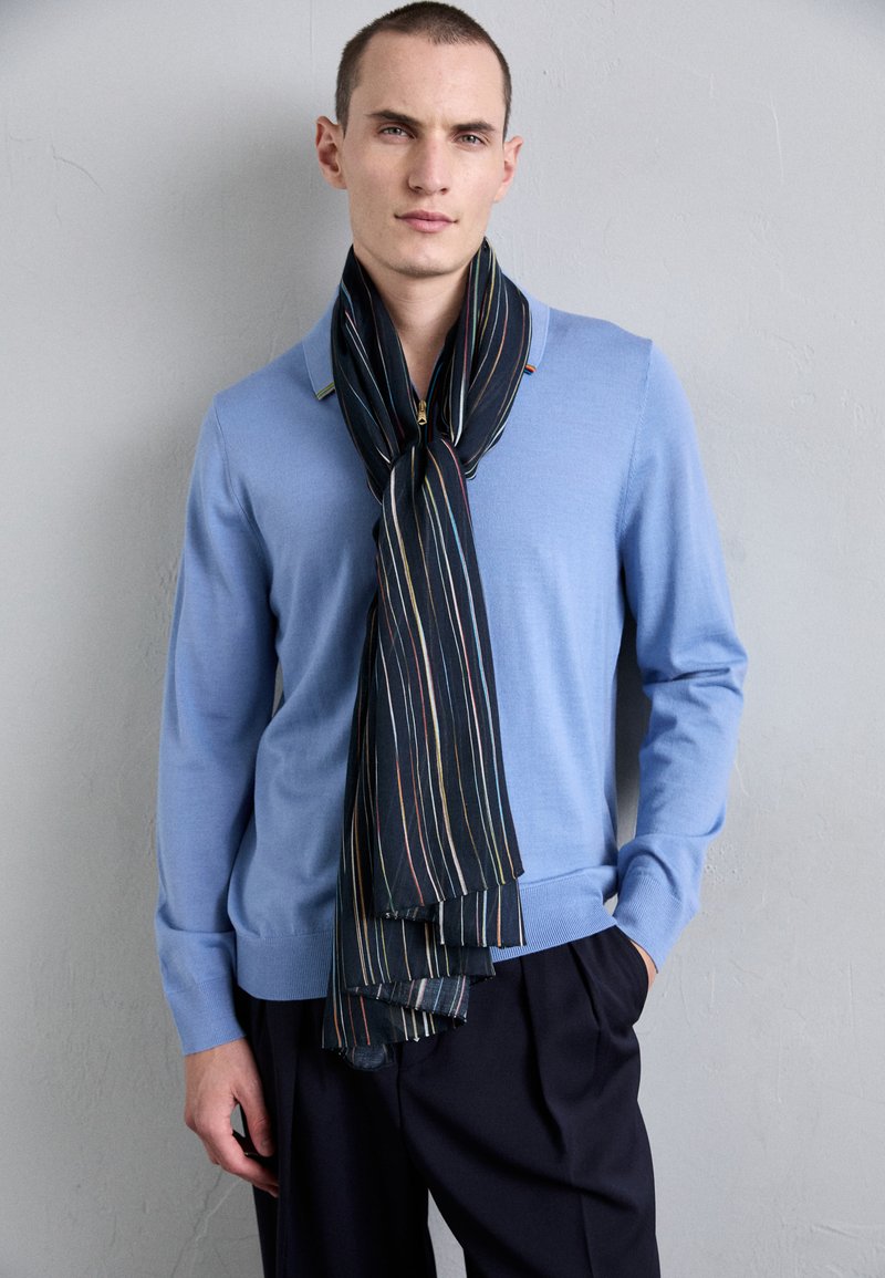 Gaiši zils džemperis ar apaļu kakla izgriezumu, apvienots ar tumši svītrainu šalli, kurai ir daudzkrāsainas svītras, eleganti valkāta ap kaklu.