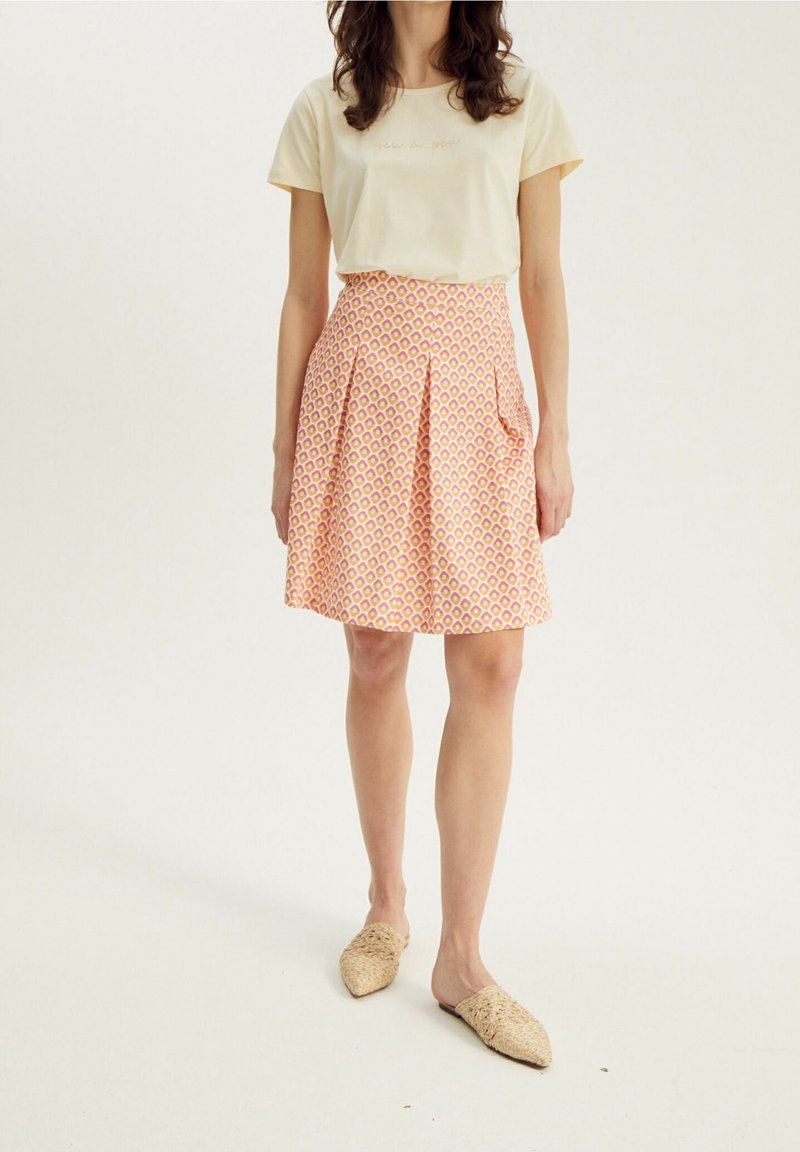 Camiseta color beige claro con texto bordado, combinada con una falda rosa con estampado que presenta pliegues y bolsillos, usada con zapatos de lona sin cordones.