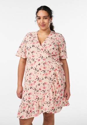 Kobieta z kręconymi włosami w różowej sukience w kwiaty, z krótkimi rękawami i falbaną u dołu, stojąca na tle jednolitego białego tła.