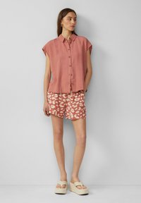Rosa Leinen-Hemd mit kurzen Ärmeln und Knöpfen, kombiniert mit hellkorallenfarbigen gemusterten Shorts. Das Modell trägt cremefarbene Plateausandalen. Neutraler Hintergrund.