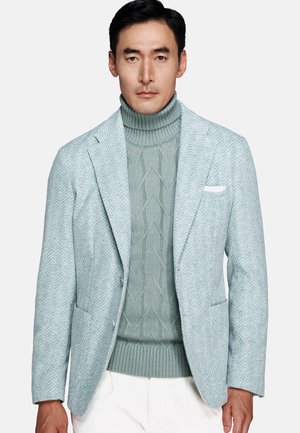 Man wearing light blue herringbone blazer over matching cable-knit turtleneck sweater and white pants against plain background.