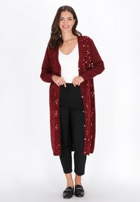 Femeie zâmbitoare îmbrăcată într-un top alb, un cardigan lung din sequins burgund, pantaloni scurți negri și mocasini negri, pe un fundal alb.