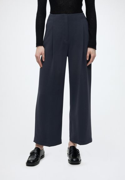Mujer con pantalones azul marino de tiro alto y pierna ancha, top de manga larga negro con ribetes y mocasines de cuero negro, de pie sobre fondo blanco.