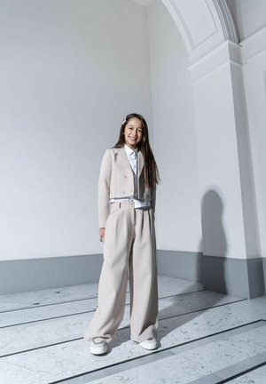 Beżowy garnitur z dopasowaną kurtką, szerokimi spodniami i białą koszulą. Wyjątkowa teksturowana tkanina i srebrne sneakersy. Prosty dodatek do włosów.