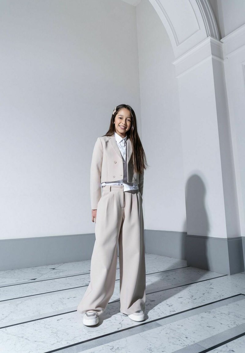 Beżowy garnitur z dopasowaną kurtką, szerokimi spodniami i białą koszulą. Wyjątkowa teksturowana tkanina i srebrne sneakersy. Prosty dodatek do włosów.