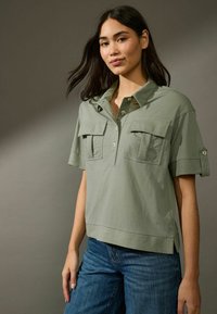 Camicetta khaki a maniche corte con colletto, due tasche sul petto e dettagli con bottoni. Abbinata a jeans in denim blu. Vestibilità morbida e rilassata.