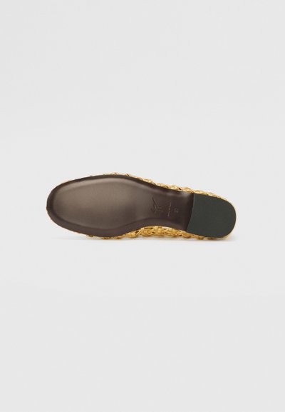 Platt sandal med en flätad guldupp, en slät mörkbrun sula, och en grön accent på hälen. Texturerad yta med vävda detaljer.